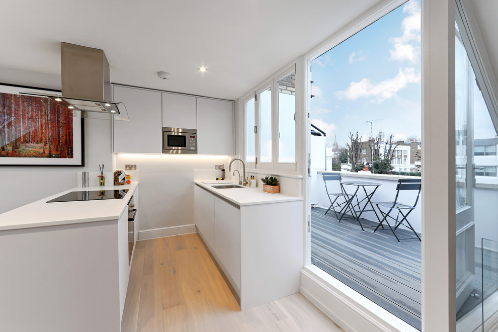 Small contemporary galley kitchen in London with a submerged sink, flat-panel cabinets, white cabinets, stainless steel appliances, beige floors, white worktops, white splashback, window splashback, light hardwood flooring and a breakfast bar.