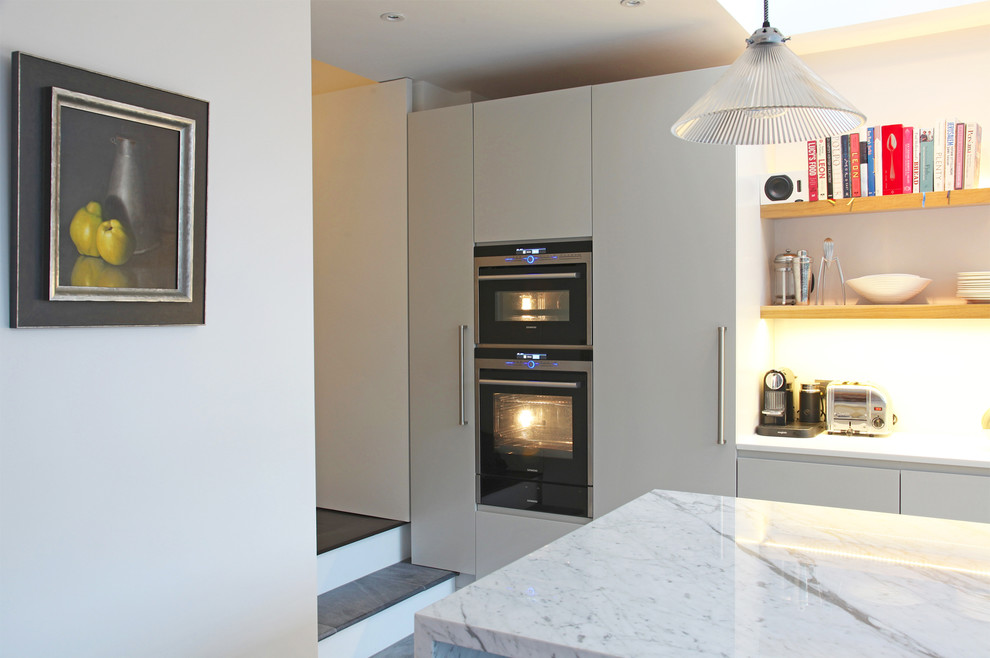 Photo of a large contemporary galley open plan kitchen in London with an integrated sink, flat-panel cabinets, grey cabinets, marble worktops, white splashback, glass sheet splashback, black appliances, ceramic flooring and an island.