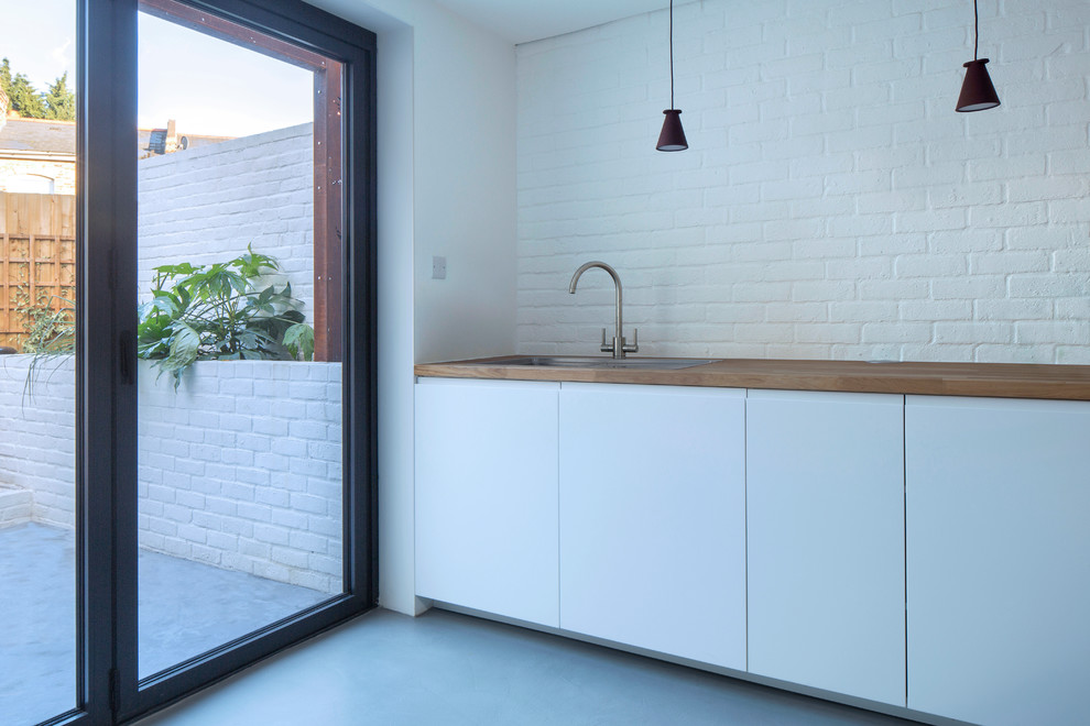 Foto på ett litet funkis linjärt kök med öppen planlösning, med en undermonterad diskho, släta luckor, vita skåp, träbänkskiva, vitt stänkskydd, stänkskydd i tegel, vita vitvaror, betonggolv och grått golv