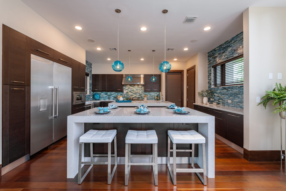 Küche in L-Form mit zwei Kücheninseln, flächenbündigen Schrankfronten, dunklen Holzschränken, Küchenrückwand in Blau, Rückwand aus Stäbchenfliesen, Küchengeräten aus Edelstahl, braunem Holzboden und weißer Arbeitsplatte in Miami