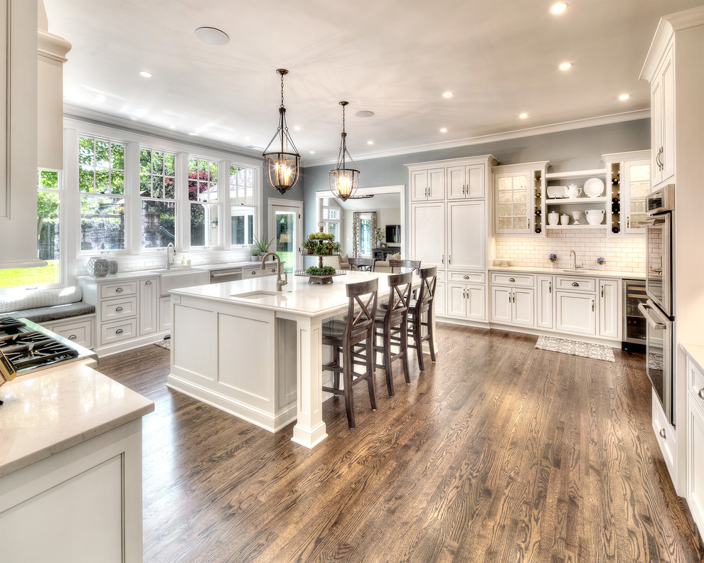 Design ideas for an expansive traditional kitchen in Kansas City with a belfast sink, recessed-panel cabinets, white cabinets, composite countertops, white splashback, stainless steel appliances, medium hardwood flooring, an island and metro tiled splashback.