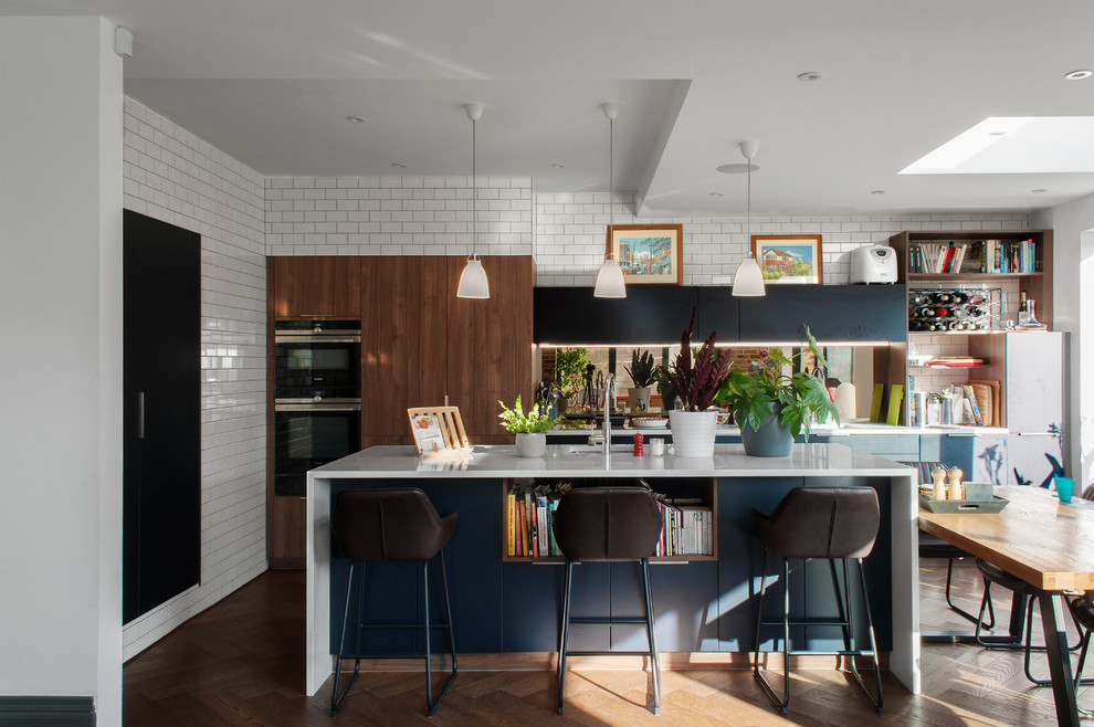 Design ideas for a medium sized contemporary l-shaped open plan kitchen in London with flat-panel cabinets, composite countertops, medium hardwood flooring, an island, white worktops, a submerged sink, medium wood cabinets, brown floors and mirror splashback.