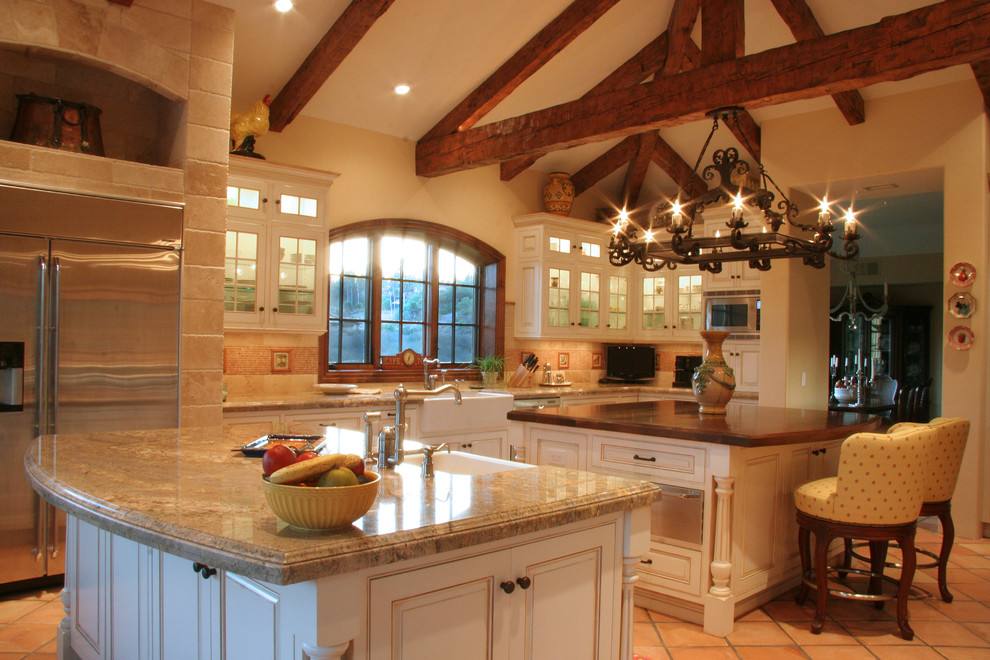 Example of a large trendy u-shaped terra-cotta tile and beige floor enclosed kitchen design in San Diego with stainless steel appliances, a farmhouse sink, raised-panel cabinets, white cabinets, granite countertops, beige backsplash, travertine backsplash, two islands and beige countertops