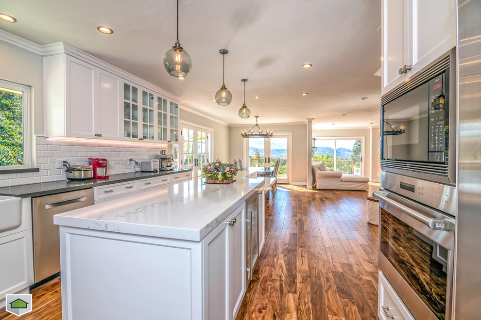 Imagen de cocina contemporánea grande con fregadero sobremueble, armarios estilo shaker, puertas de armario blancas, encimera de mármol, salpicadero blanco, salpicadero de azulejos tipo metro, electrodomésticos de acero inoxidable, suelo de madera en tonos medios y una isla