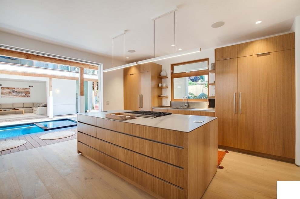 Photo of a large world-inspired galley open plan kitchen in Los Angeles with a submerged sink, flat-panel cabinets, medium wood cabinets, engineered stone countertops, metallic splashback, window splashback, stainless steel appliances, light hardwood flooring, an island, brown floors and grey worktops.