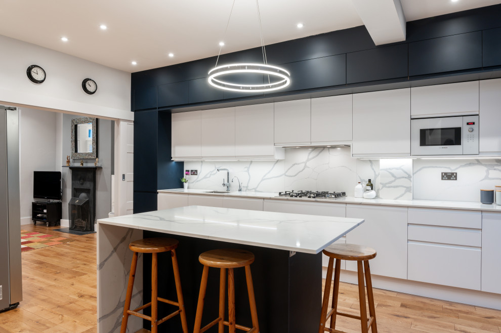 Offene Moderne Küche mit flächenbündigen Schrankfronten, blauen Schränken, Quarzit-Arbeitsplatte, Küchenrückwand in Weiß, Elektrogeräten mit Frontblende, braunem Holzboden, Kücheninsel und weißer Arbeitsplatte in London