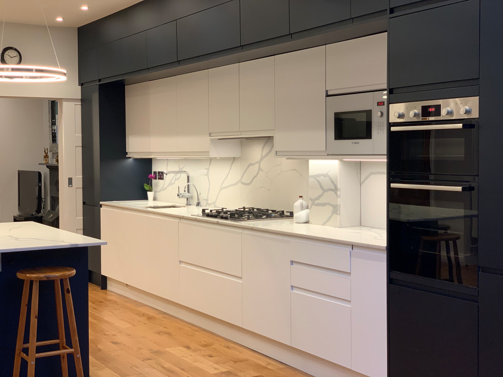 Cette photo montre une cuisine ouverte encastrable moderne avec un placard à porte plane, des portes de placard bleues, un plan de travail en quartz, une crédence blanche, un sol en bois brun, îlot et un plan de travail blanc.