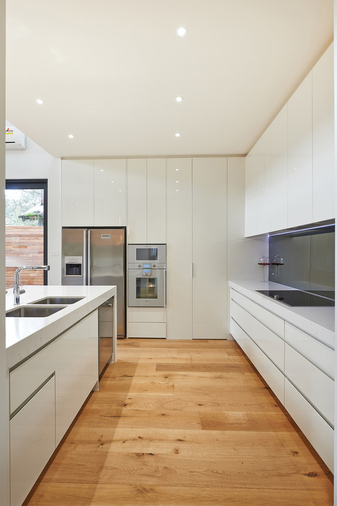 Mittelgroße Moderne Wohnküche mit Doppelwaschbecken, flächenbündigen Schrankfronten, weißen Schränken, Quarzwerkstein-Arbeitsplatte, Küchenrückwand in Grau, Glasrückwand, Küchengeräten aus Edelstahl, braunem Holzboden und Kücheninsel in Melbourne