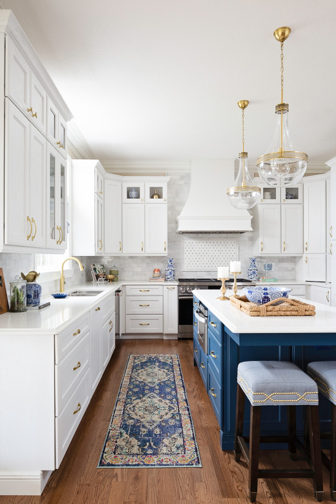 Klassisk inredning av ett vit vitt l-kök, med en undermonterad diskho, skåp i shakerstil, vita skåp, grått stänkskydd, rostfria vitvaror, mellanmörkt trägolv, en köksö och brunt golv