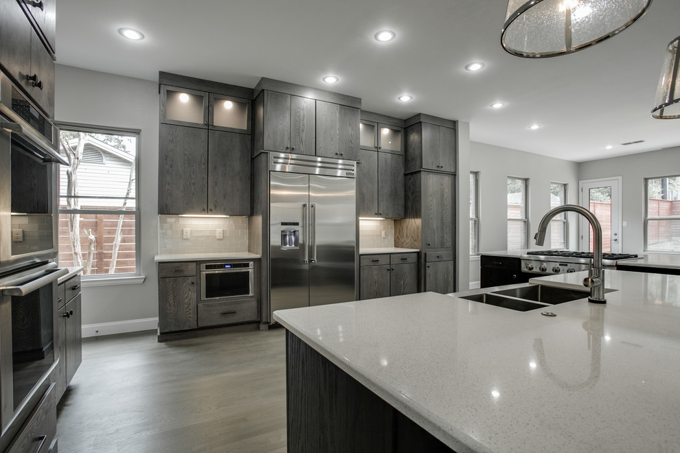 This is an example of a large modern u-shaped open plan kitchen in Dallas with a belfast sink, flat-panel cabinets, grey cabinets, engineered stone countertops, white splashback, metro tiled splashback, stainless steel appliances, light hardwood flooring and multiple islands.