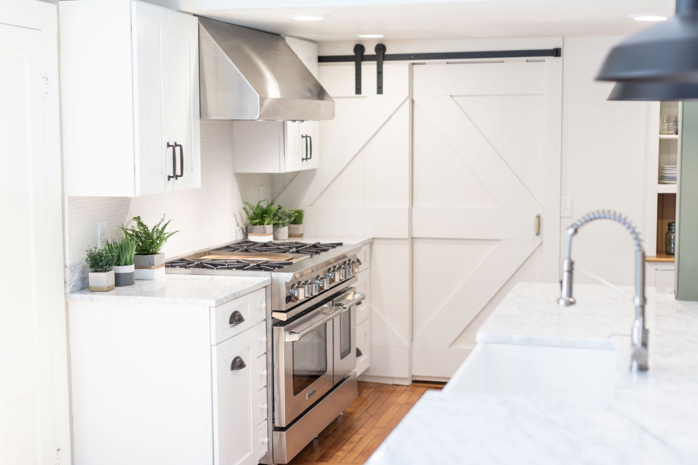 Photo of a large country galley kitchen/diner in Burlington with a belfast sink, shaker cabinets, white cabinets, marble worktops, white splashback, metro tiled splashback, stainless steel appliances, light hardwood flooring, an island and white worktops.