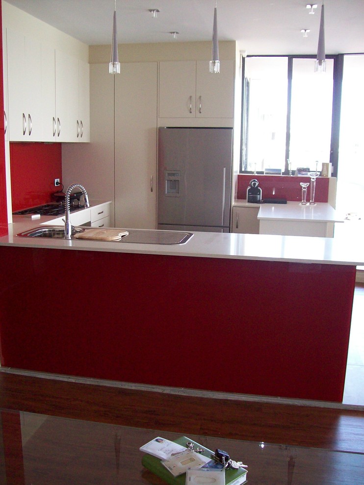 Photo of a medium sized modern u-shaped kitchen in Sydney with a built-in sink, white cabinets, engineered stone countertops, red splashback, glass sheet splashback, stainless steel appliances, porcelain flooring and a breakfast bar.