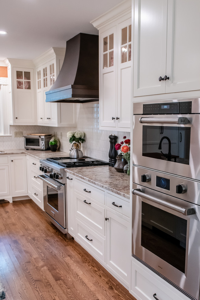 Mid-sized elegant l-shaped medium tone wood floor and brown floor enclosed kitchen photo in Portland Maine with an undermount sink, recessed-panel cabinets, white cabinets, granite countertops, white backsplash, paneled appliances, an island and beige countertops
