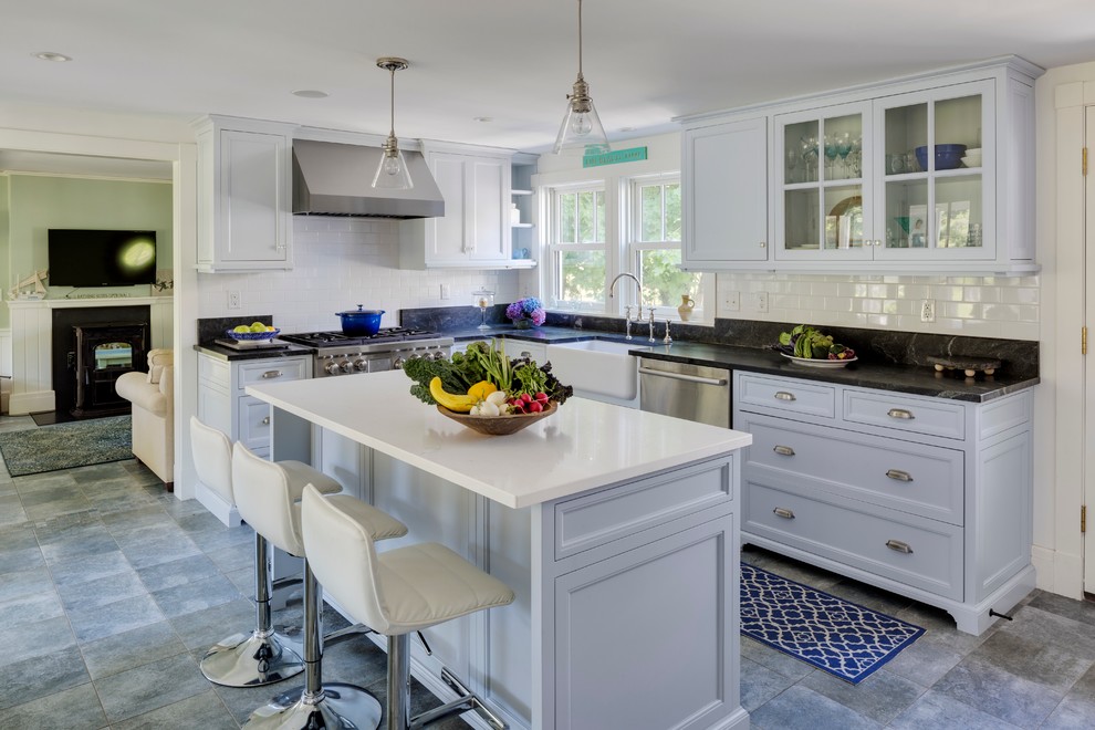 Medium sized nautical l-shaped kitchen/diner in Boston with white cabinets, an island, a belfast sink, glass-front cabinets, white splashback, stainless steel appliances and grey floors.
