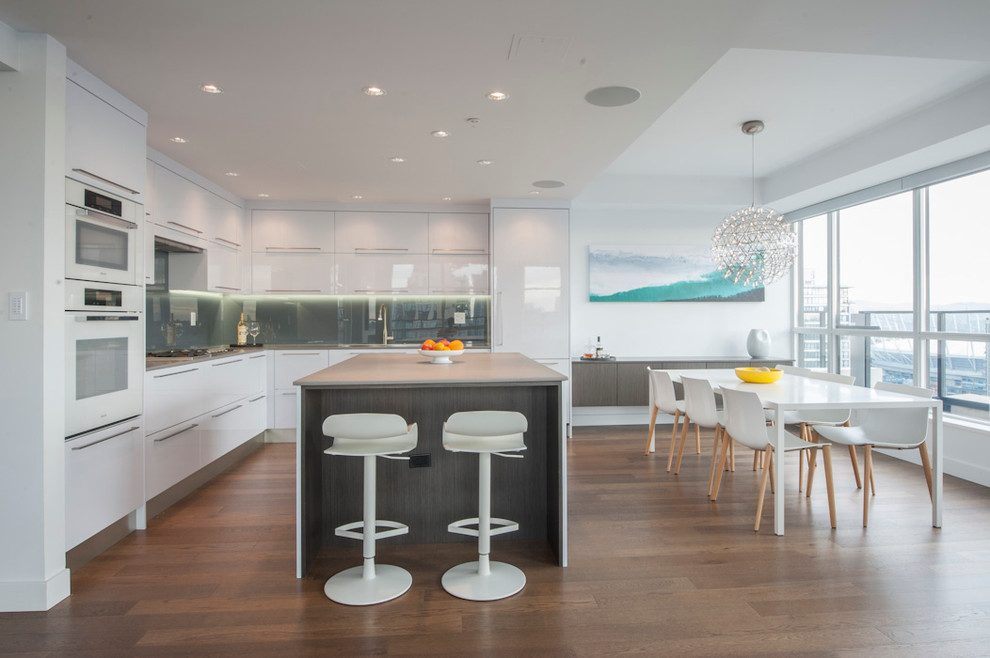 Trendy l-shaped medium tone wood floor eat-in kitchen photo in Vancouver with flat-panel cabinets, white cabinets, gray backsplash, glass sheet backsplash, white appliances and an island