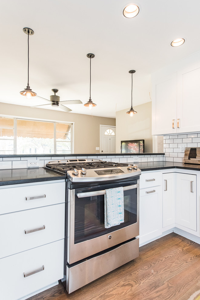 Example of a large classic l-shaped medium tone wood floor eat-in kitchen design in Birmingham with a farmhouse sink, shaker cabinets, white cabinets, granite countertops, white backsplash, ceramic backsplash, stainless steel appliances and a peninsula