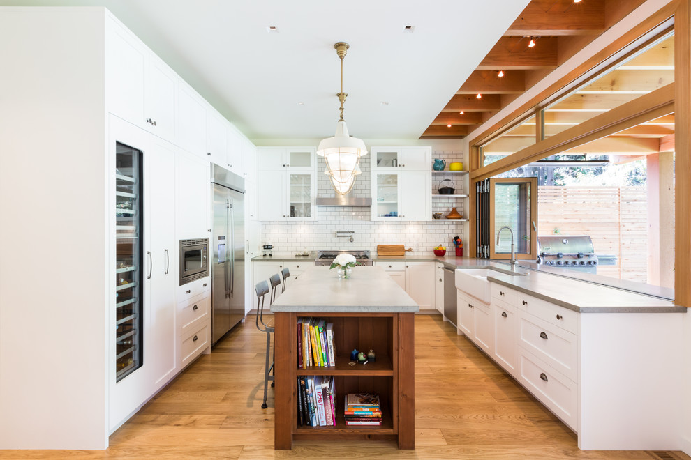 Inspiration for a large contemporary u-shaped kitchen/diner in Vancouver with white cabinets, concrete worktops, white splashback, metro tiled splashback, stainless steel appliances, an island, a belfast sink, shaker cabinets, brown floors, medium hardwood flooring and grey worktops.