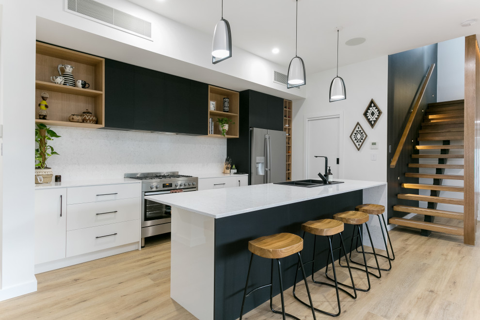Photo of a large contemporary galley open plan kitchen in Adelaide with a built-in sink, white cabinets, engineered stone countertops, white splashback, stone slab splashback, stainless steel appliances, light hardwood flooring, an island, multi-coloured floors and white worktops.