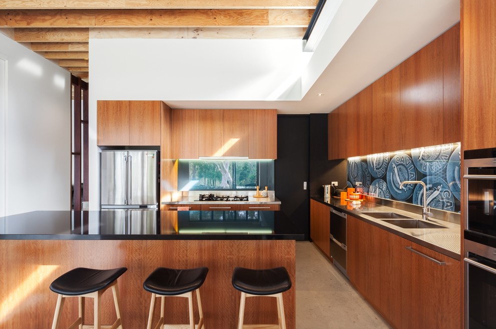 Foto på ett funkis l-kök, med en dubbel diskho, släta luckor, skåp i mellenmörkt trä, bänkskiva i rostfritt stål, blått stänkskydd, rostfria vitvaror och en köksö