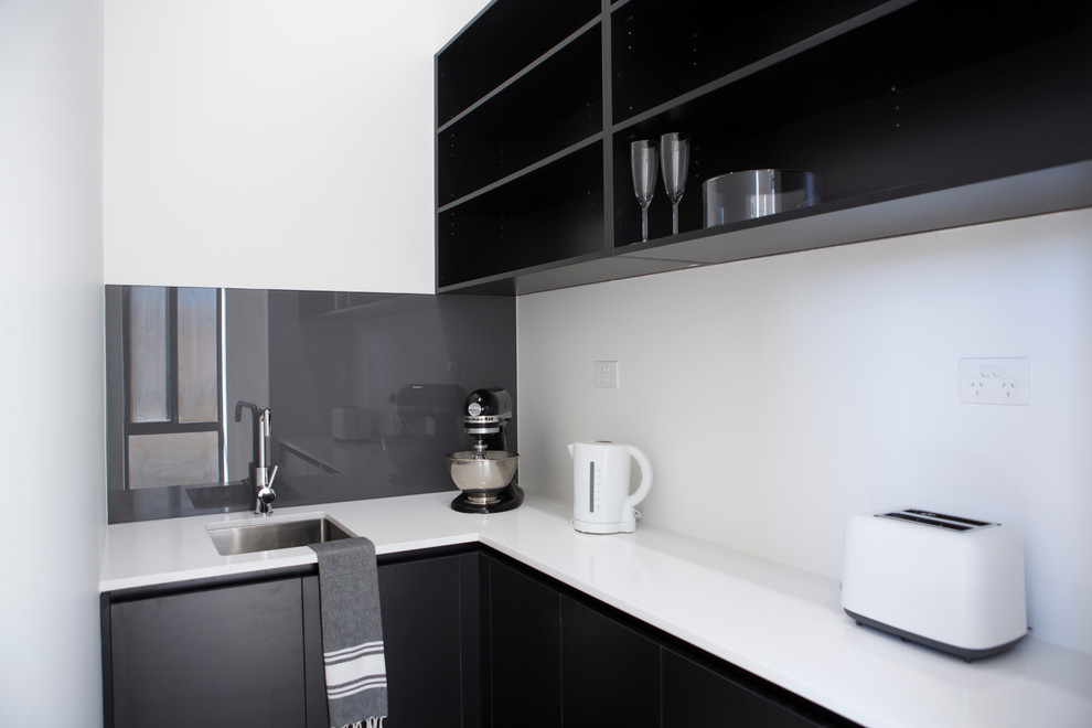 Inspiration for a medium sized contemporary l-shaped kitchen pantry in Melbourne with black cabinets, engineered stone countertops, grey splashback, glass sheet splashback, concrete flooring, a single-bowl sink, flat-panel cabinets, white appliances and no island.