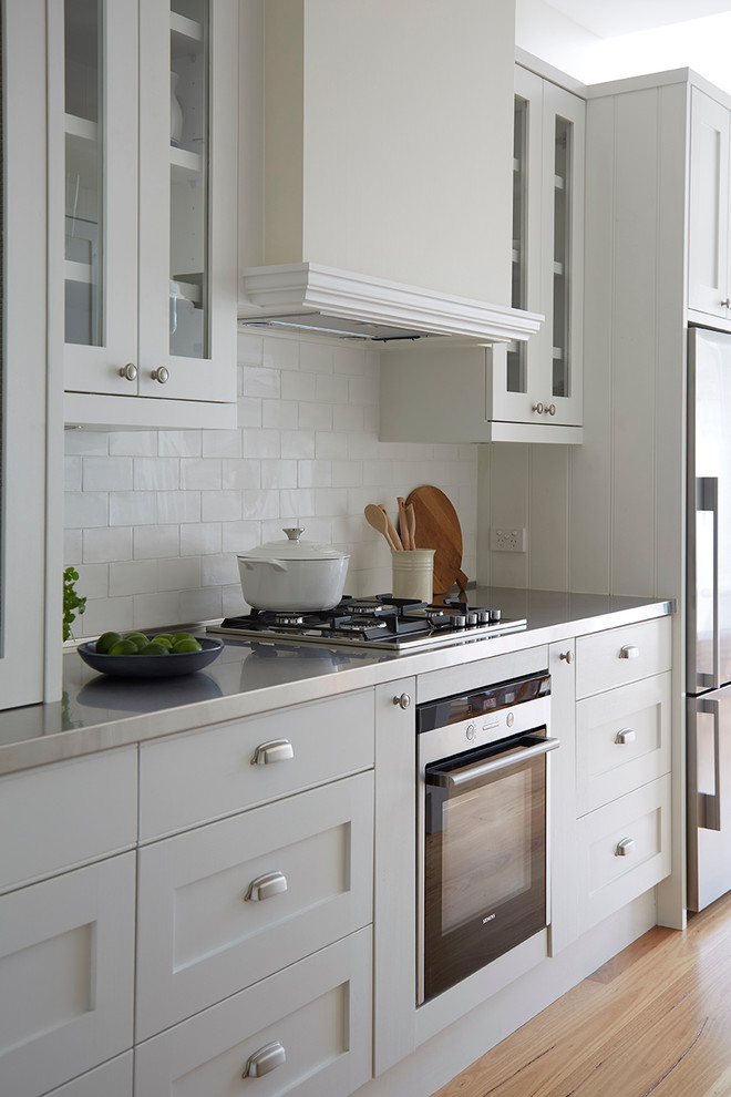 Klassisk inredning av ett mellanstort kök, med en undermonterad diskho, skåp i shakerstil, vita skåp, marmorbänkskiva, vitt stänkskydd, stänkskydd i porslinskakel, rostfria vitvaror, ljust trägolv och en köksö