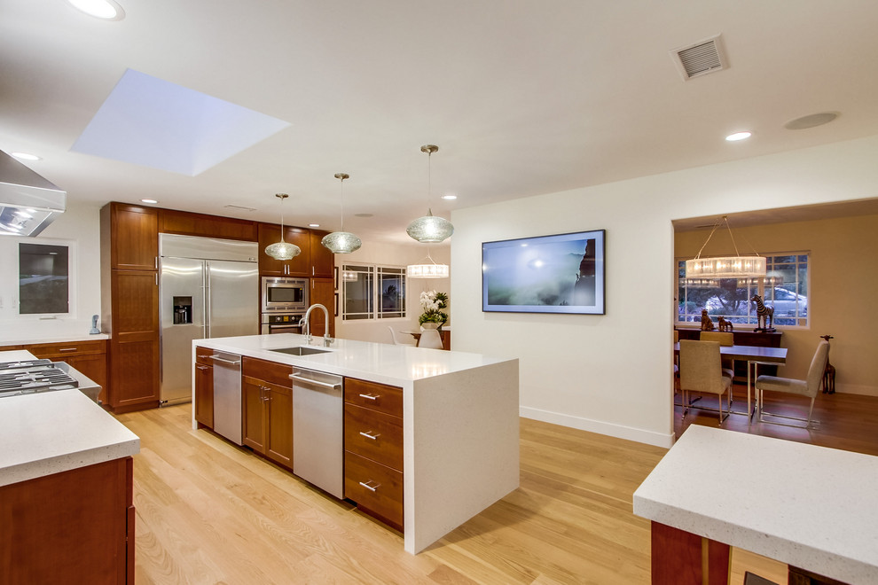 Large midcentury l-shaped enclosed kitchen with a single-bowl sink, recessed-panel cabinets, medium wood cabinets, engineered stone countertops, blue splashback, glass tiled splashback, stainless steel appliances, light hardwood flooring, an island, beige floors and white worktops.