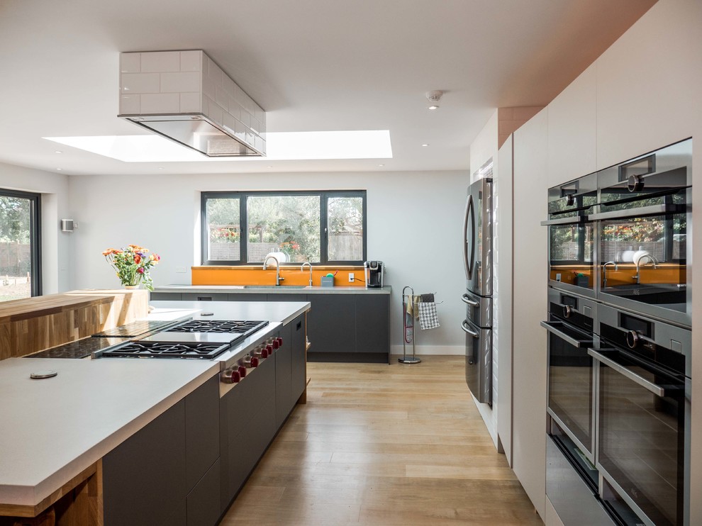 Inspiration for a large contemporary l-shaped open plan kitchen in London with an integrated sink, recessed-panel cabinets, white cabinets, concrete worktops, stainless steel appliances, medium hardwood flooring, an island, brown floors, orange splashback and glass sheet splashback.