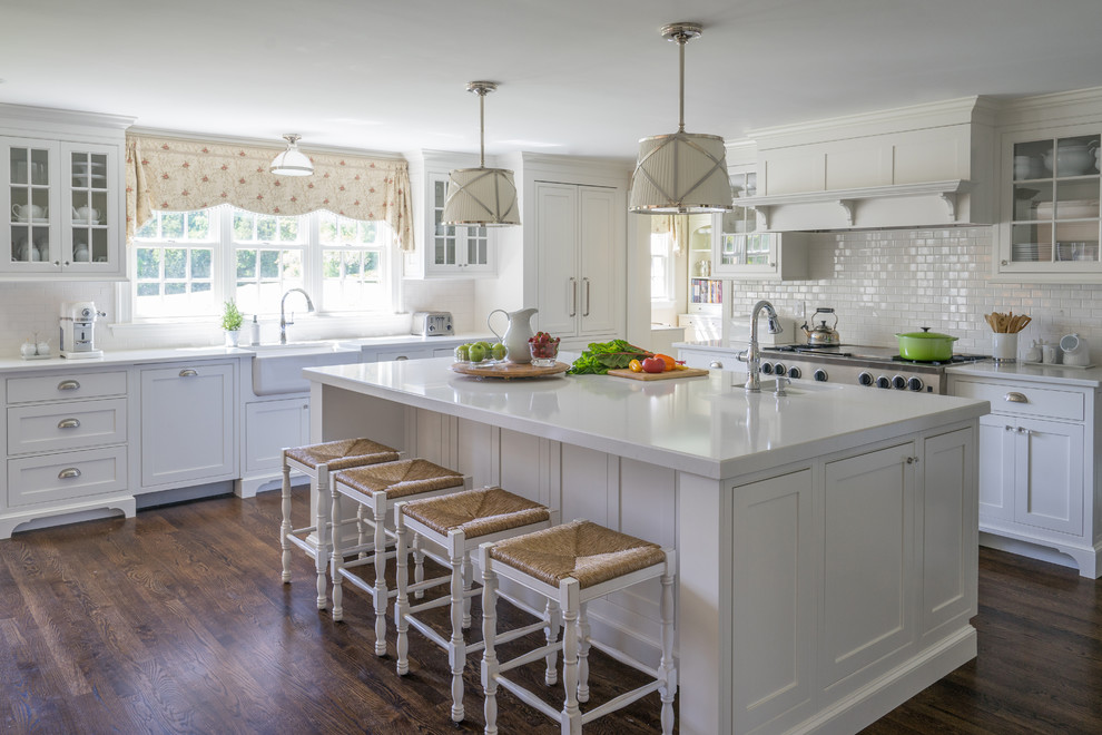 Large country l-shaped kitchen/diner in Boston with a belfast sink, recessed-panel cabinets, white cabinets, engineered stone countertops, white splashback, ceramic splashback, stainless steel appliances, medium hardwood flooring, an island and brown floors.