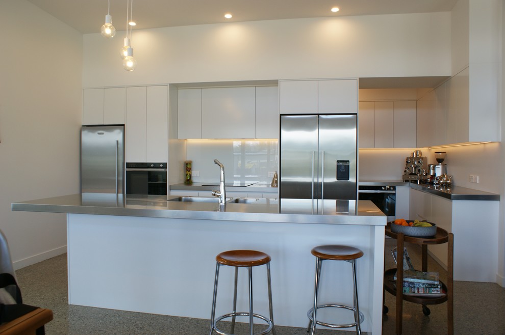 Large urban galley concrete floor kitchen pantry photo in Dunedin with an integrated sink, flat-panel cabinets, white cabinets, stainless steel countertops, white backsplash, glass sheet backsplash, stainless steel appliances and an island