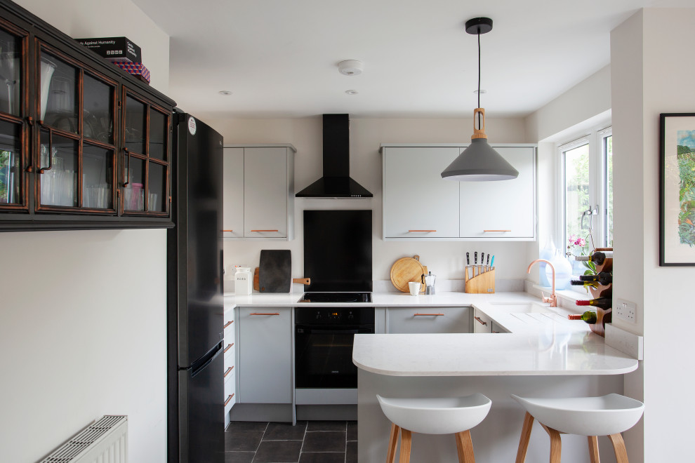 Example of a small trendy u-shaped cement tile floor and black floor open concept kitchen design in London with a drop-in sink, flat-panel cabinets, gray cabinets, solid surface countertops, white backsplash, quartz backsplash, black appliances, no island and white countertops