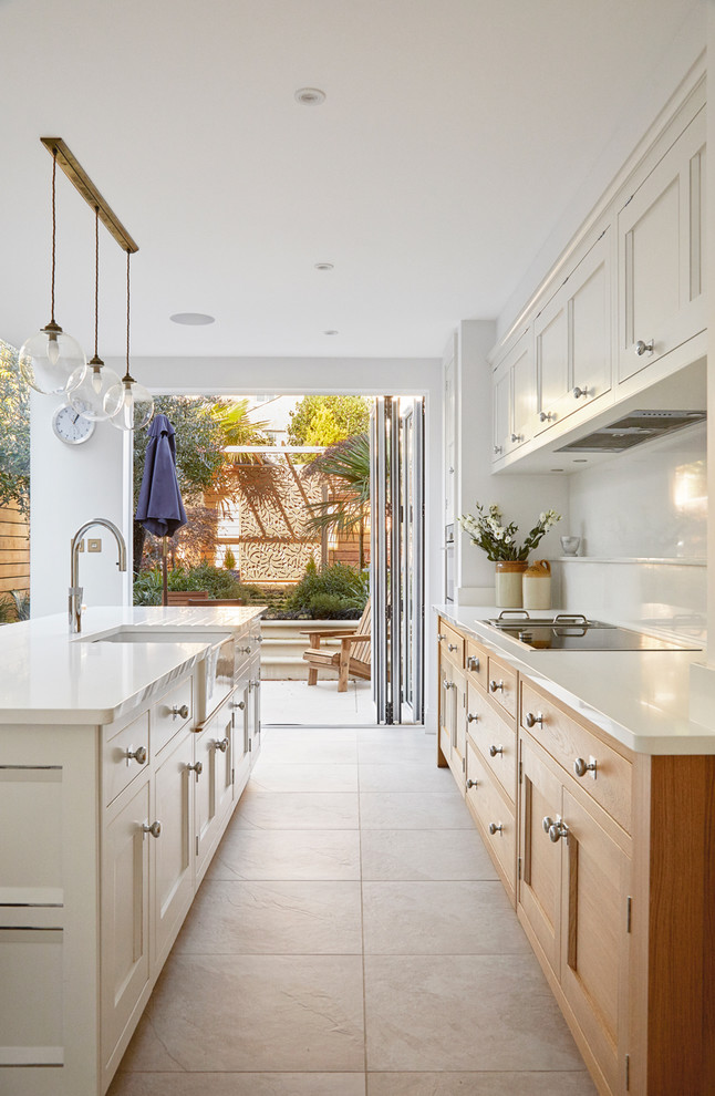 This is an example of a traditional galley kitchen in London with a belfast sink, recessed-panel cabinets, white cabinets, white splashback, black appliances, an island, beige floors and white worktops.