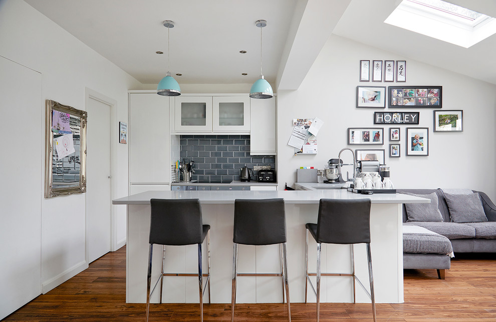 Photo of a contemporary l-shaped open plan kitchen in London with flat-panel cabinets, white cabinets, grey splashback, metro tiled splashback, medium hardwood flooring, a breakfast bar and brown floors.