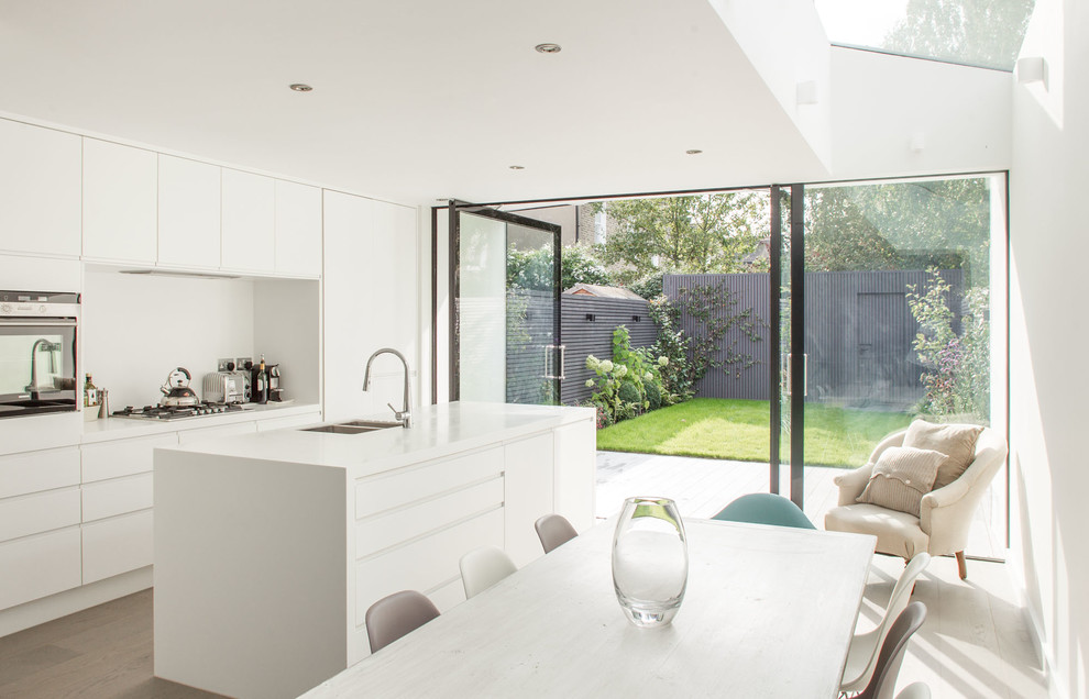 Modern inredning av ett vit vitt kök, med en undermonterad diskho, släta luckor, vita skåp, vitt stänkskydd, glaspanel som stänkskydd, mellanmörkt trägolv, en köksö, rostfria vitvaror och brunt golv