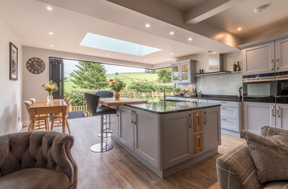 Large classic single-wall open plan kitchen in Cheshire with a submerged sink, shaker cabinets, blue cabinets, granite worktops, white splashback, glass sheet splashback, black appliances, vinyl flooring, an island and beige floors.