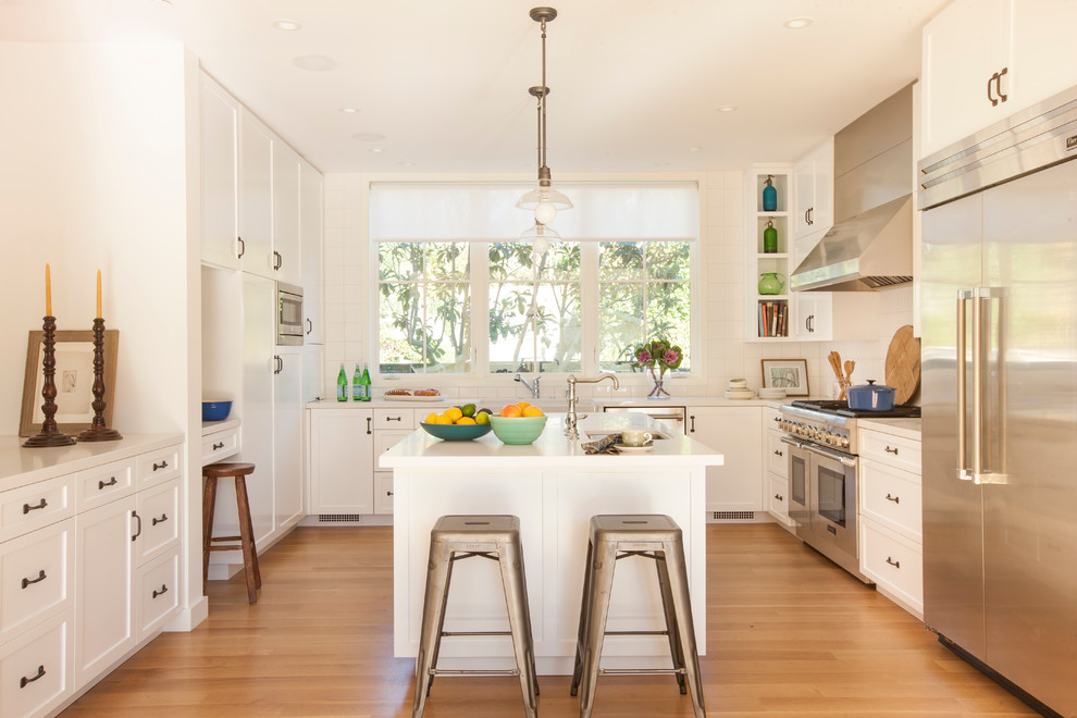 Enclosed kitchen - large transitional u-shaped light wood floor and brown floor enclosed kitchen idea in Los Angeles with a farmhouse sink, shaker cabinets, white cabinets, solid surface countertops, white backsplash, porcelain backsplash, stainless steel appliances and an island