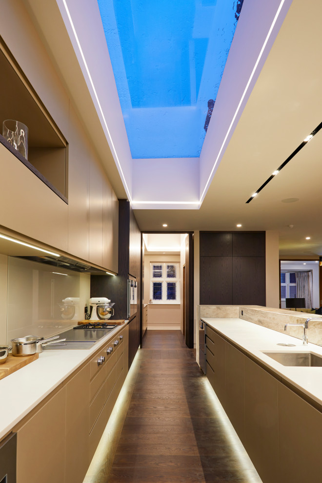Large contemporary galley kitchen in London with a submerged sink, flat-panel cabinets, composite countertops, beige splashback, glass tiled splashback, integrated appliances, dark hardwood flooring, brown floors, white worktops, beige cabinets and no island.