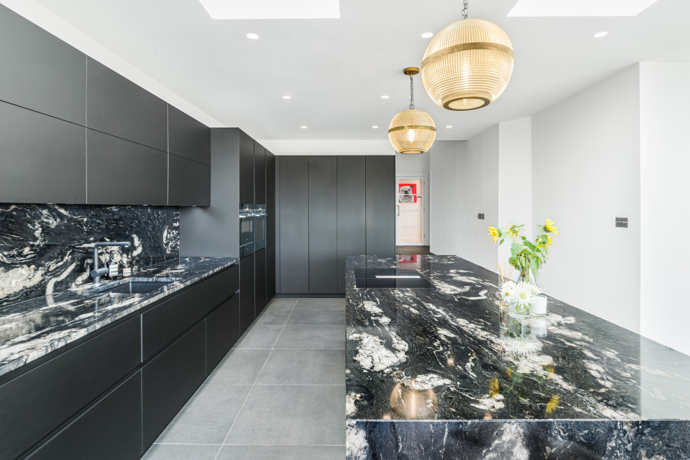 Large contemporary l-shaped open plan kitchen in Sussex with an integrated sink, flat-panel cabinets, black cabinets, granite worktops, black splashback, granite splashback, black appliances, an island, grey floors and black worktops.