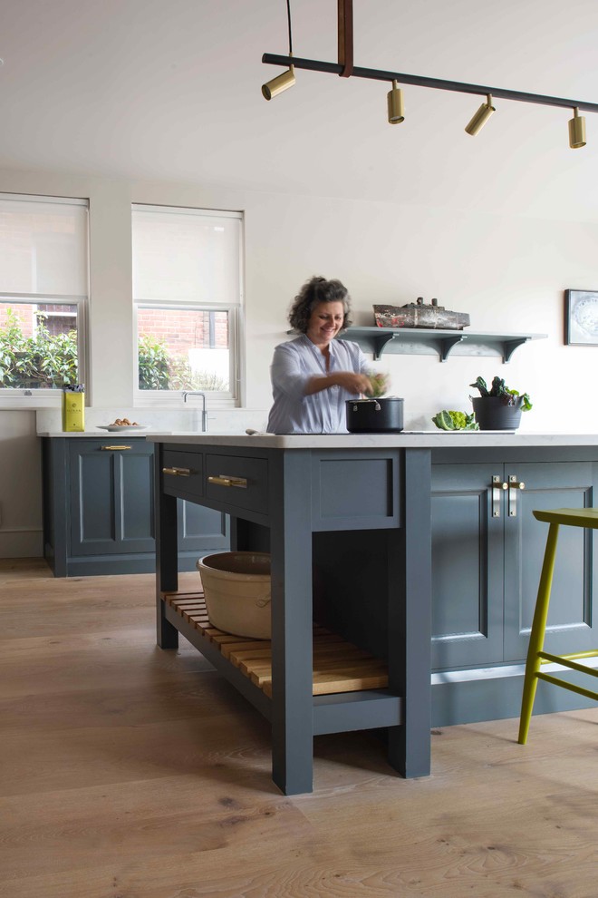 Photo of a medium sized traditional l-shaped open plan kitchen in Sussex with a belfast sink, beaded cabinets, blue cabinets, composite countertops, light hardwood flooring, an island and white worktops.