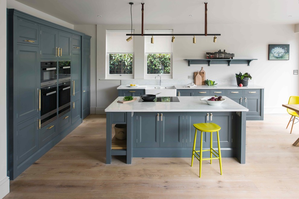 Design ideas for a medium sized classic l-shaped open plan kitchen in Sussex with a belfast sink, beaded cabinets, blue cabinets, composite countertops, light hardwood flooring, an island and white worktops.
