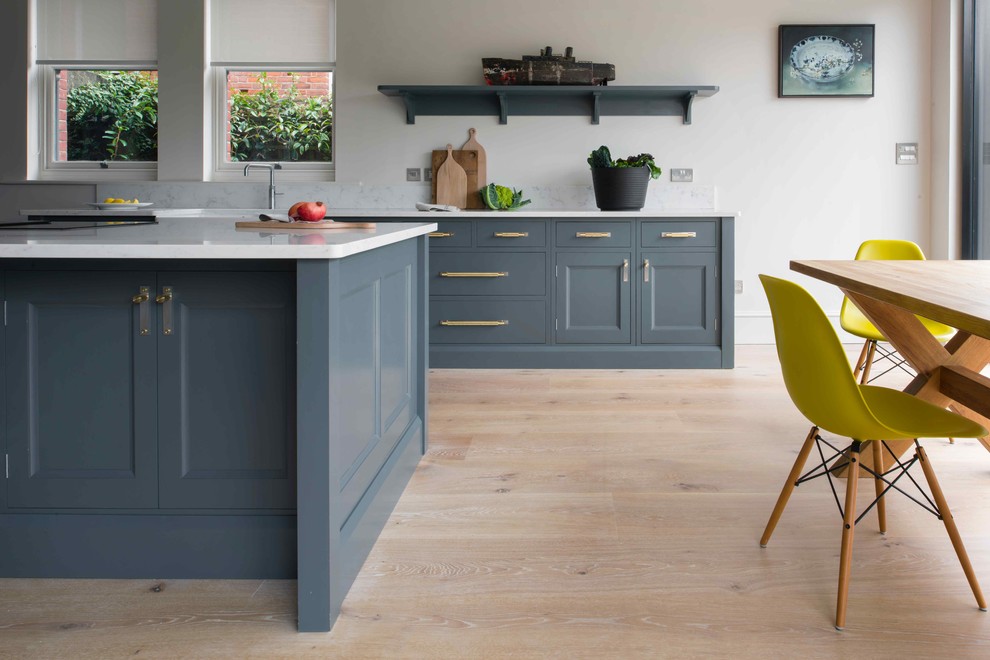 Medium sized traditional l-shaped open plan kitchen in Sussex with a belfast sink, beaded cabinets, blue cabinets, composite countertops, light hardwood flooring, an island and white worktops.