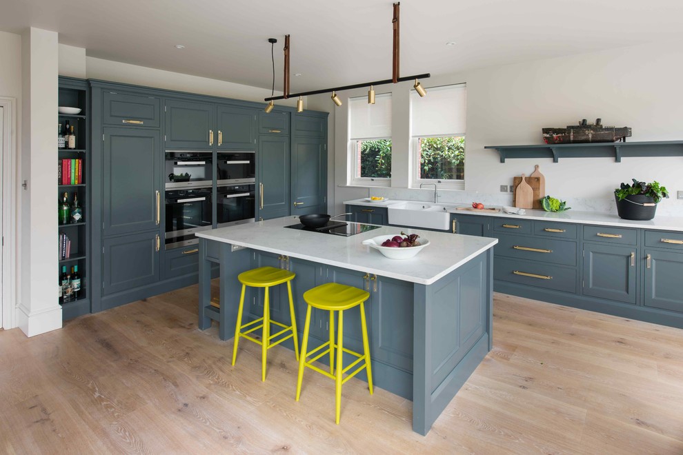 Inspiration for a medium sized traditional l-shaped open plan kitchen in Sussex with a belfast sink, beaded cabinets, blue cabinets, composite countertops, light hardwood flooring, an island and white worktops.