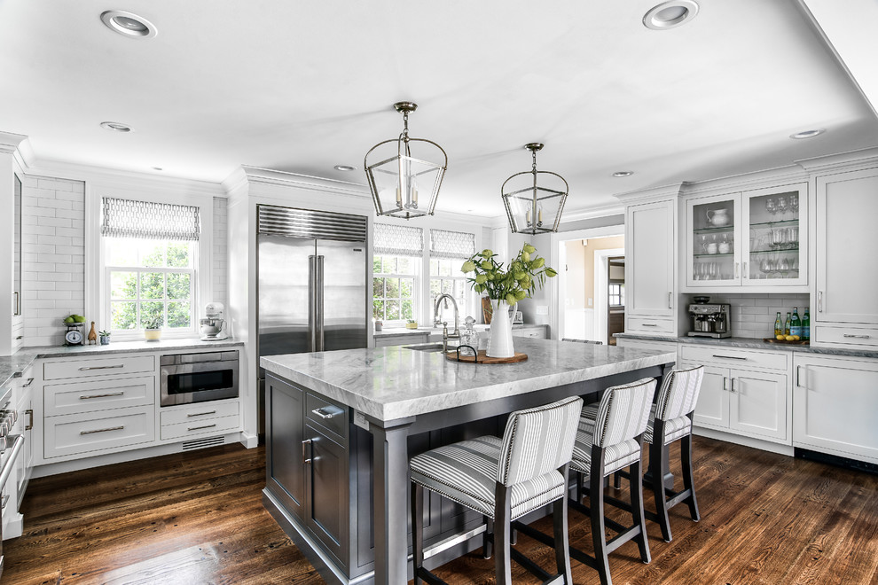 Inspiration for a transitional u-shaped dark wood floor and brown floor kitchen remodel in New York with an undermount sink, shaker cabinets, white cabinets, white backsplash, subway tile backsplash, stainless steel appliances, an island and gray countertops