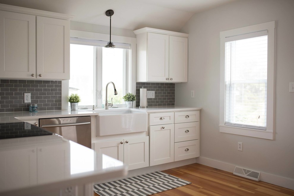 Inspiration for a medium sized nautical l-shaped kitchen/diner in Providence with a belfast sink, shaker cabinets, white cabinets, engineered stone countertops, grey splashback, metro tiled splashback, stainless steel appliances, light hardwood flooring and a breakfast bar.