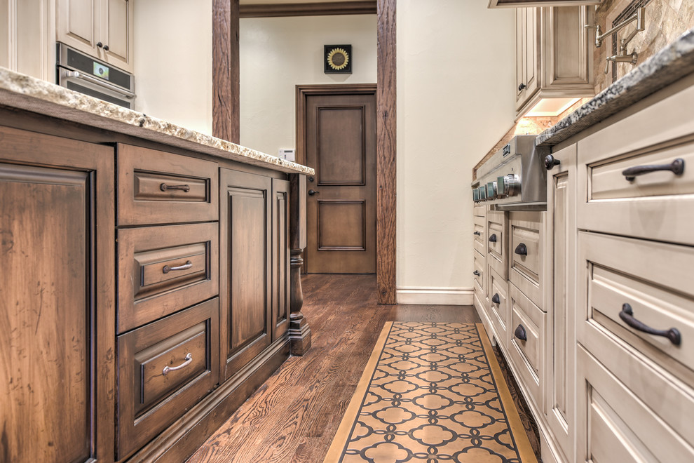 Design ideas for an expansive galley enclosed kitchen in Oklahoma City with a belfast sink, raised-panel cabinets, light wood cabinets, granite worktops, brown splashback, travertine splashback, stainless steel appliances, dark hardwood flooring, an island and brown floors.