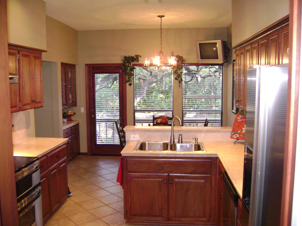 Example of a mid-sized trendy u-shaped porcelain tile kitchen design in Austin with a drop-in sink, raised-panel cabinets, medium tone wood cabinets, tile countertops, beige backsplash, stone tile backsplash, stainless steel appliances and a peninsula