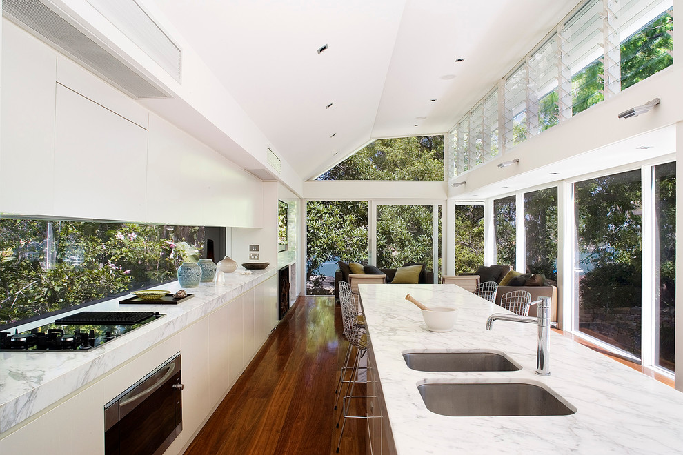Inredning av ett modernt mellanstort linjärt kök med öppen planlösning, med en dubbel diskho, släta luckor, vita skåp, marmorbänkskiva, fönster som stänkskydd, rostfria vitvaror, mellanmörkt trägolv och en köksö