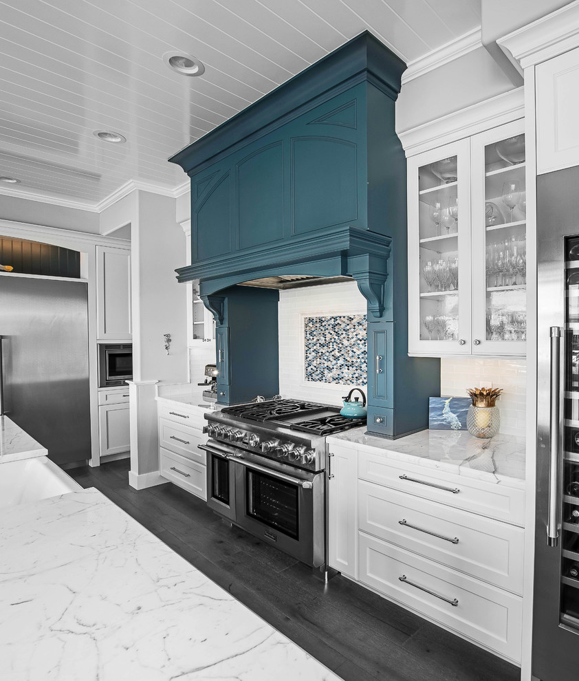 Inredning av ett maritimt stort vit vitt kök, med en rustik diskho, skåp i shakerstil, vita skåp, bänkskiva i kvarts, vitt stänkskydd, stänkskydd i porslinskakel, rostfria vitvaror, mellanmörkt trägolv, en köksö och grått golv