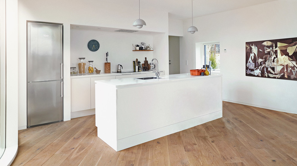 Inspiration for a medium sized scandi single-wall kitchen in Copenhagen with a built-in sink, flat-panel cabinets, white cabinets, engineered stone countertops, stainless steel appliances, light hardwood flooring, an island and white splashback.