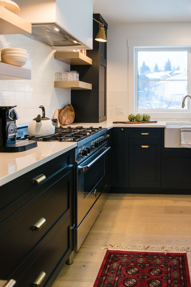 Example of a mid-sized transitional l-shaped light wood floor eat-in kitchen design in Vancouver with a farmhouse sink, shaker cabinets, black cabinets, quartzite countertops, white backsplash, ceramic backsplash, stainless steel appliances, an island and white countertops