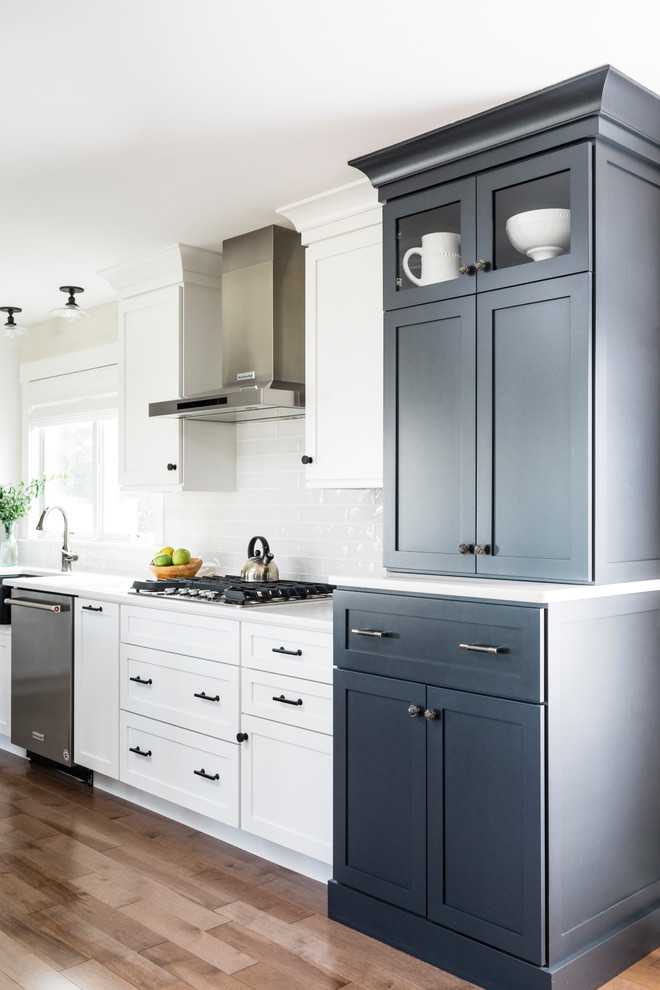 Example of a large beach style l-shaped medium tone wood floor and gray floor eat-in kitchen design in Boston with a farmhouse sink, raised-panel cabinets, blue cabinets, quartz countertops, white backsplash, glass tile backsplash, stainless steel appliances, an island and white countertops